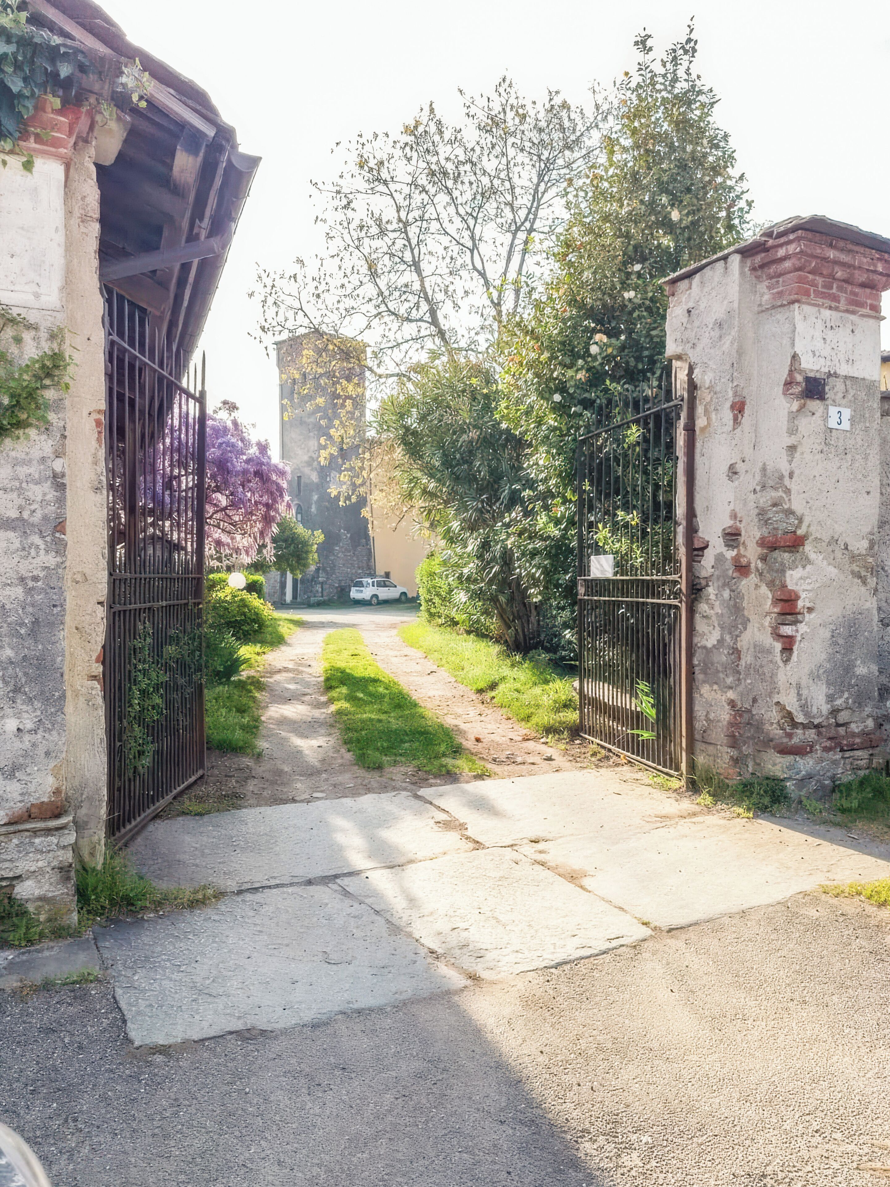 A pathway leading to a gated area with a vehicle parked in the distance, surrounded by greenery and a building with a stone facade.
