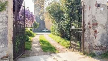 A pathway leading to a gated area with a vehicle parked in the distance, surrounded by greenery and a building with a stone facade.