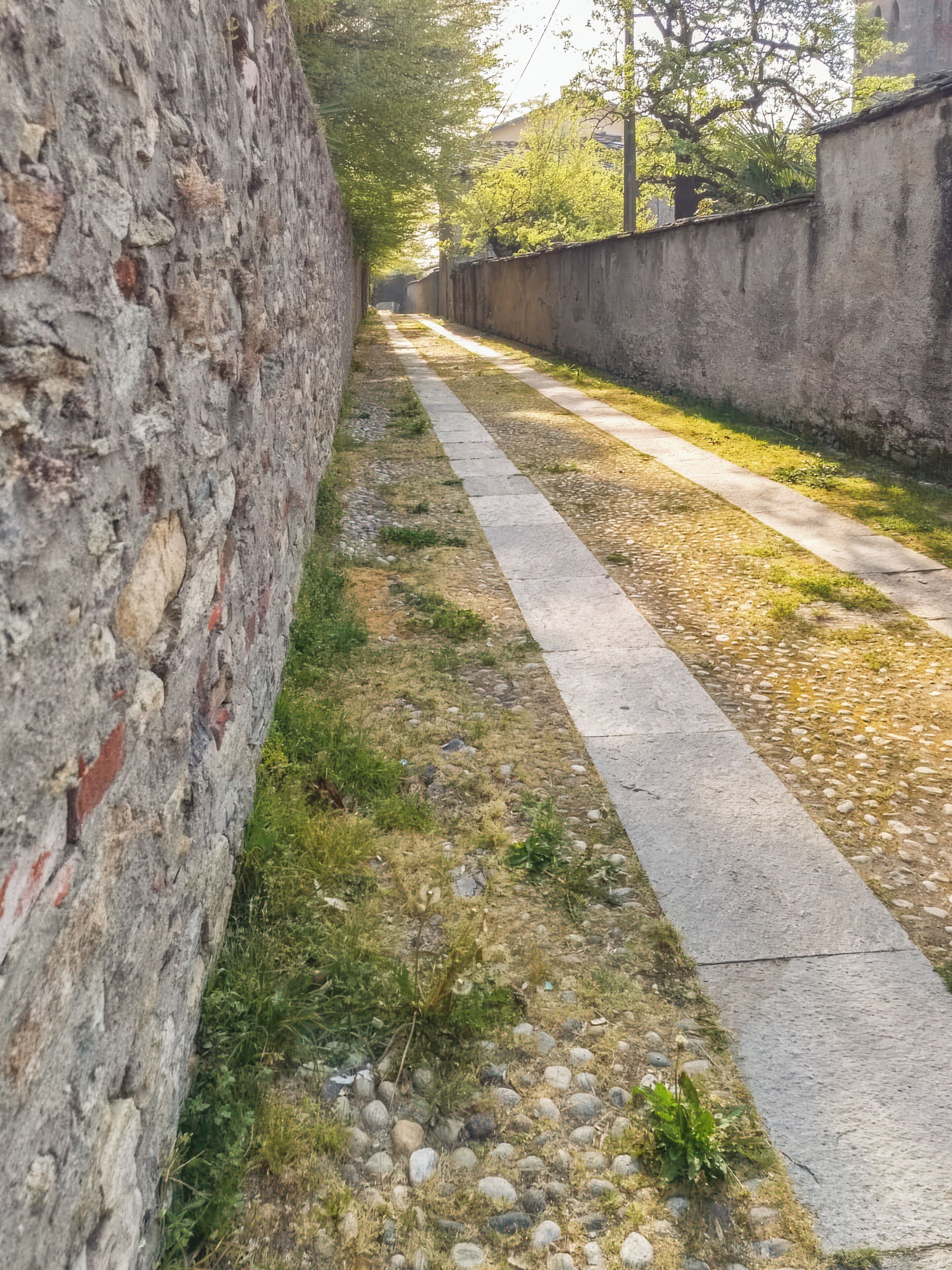 Un sentiero stretto delimitato da mura in pietra, con un percorso in cemento che costeggia.