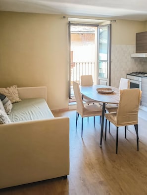 A modern living room with a sofa, dining table, and chairs, a kitchen area with a stove, and a balcony with a railing.