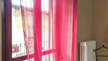 Una habitación con una ventana cubierta por cortinas delgadas color rosa, un radiador y un perchero metálico.