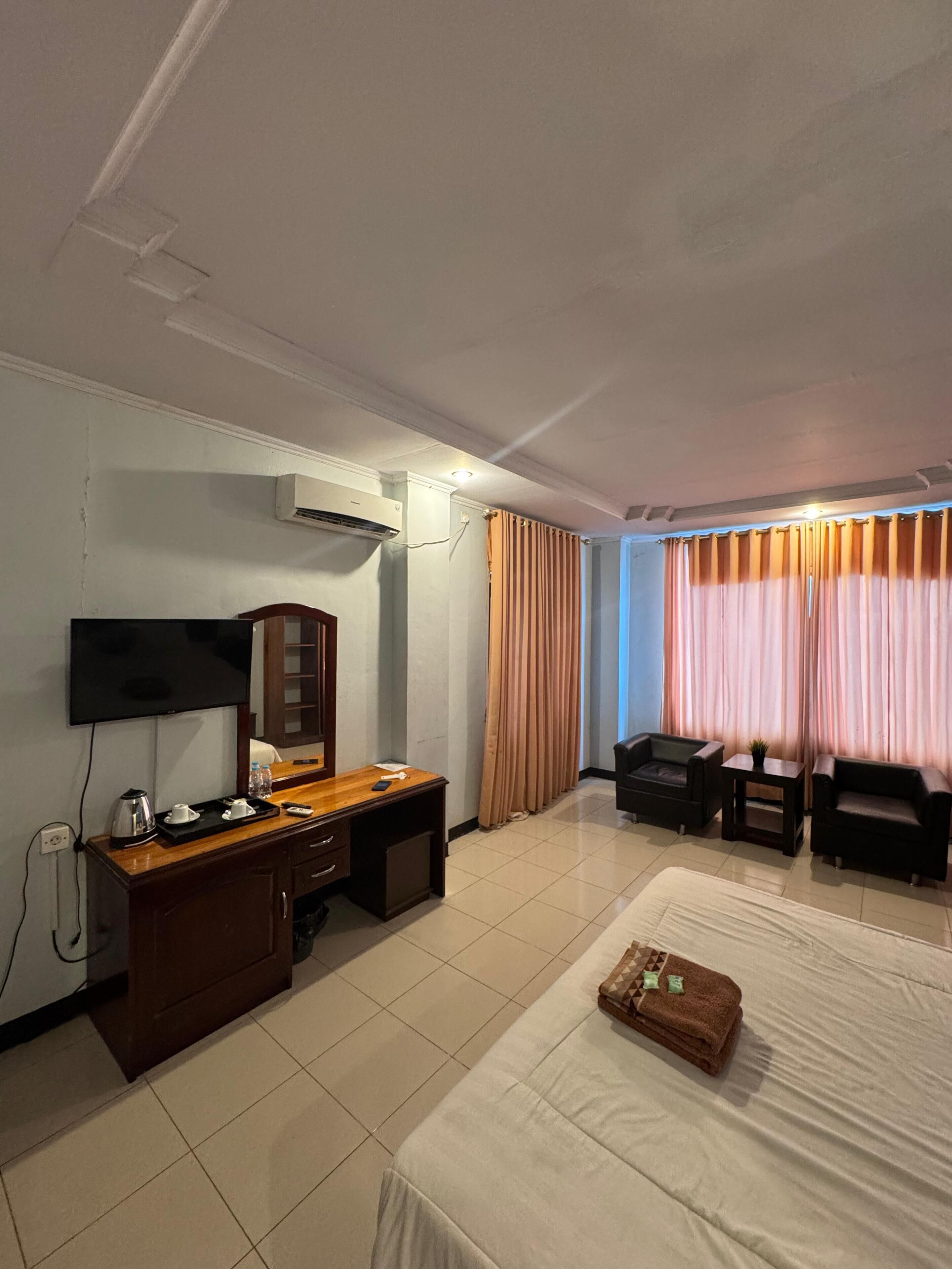 A hotel room with a bed, a desk with a flat-screen TV, a wardrobe, a small table with a plant, and a window with curtains.