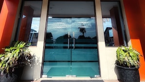 Entrada de un edificio con puerta de cristal, dos plantas en macetas y un felpudo negro.