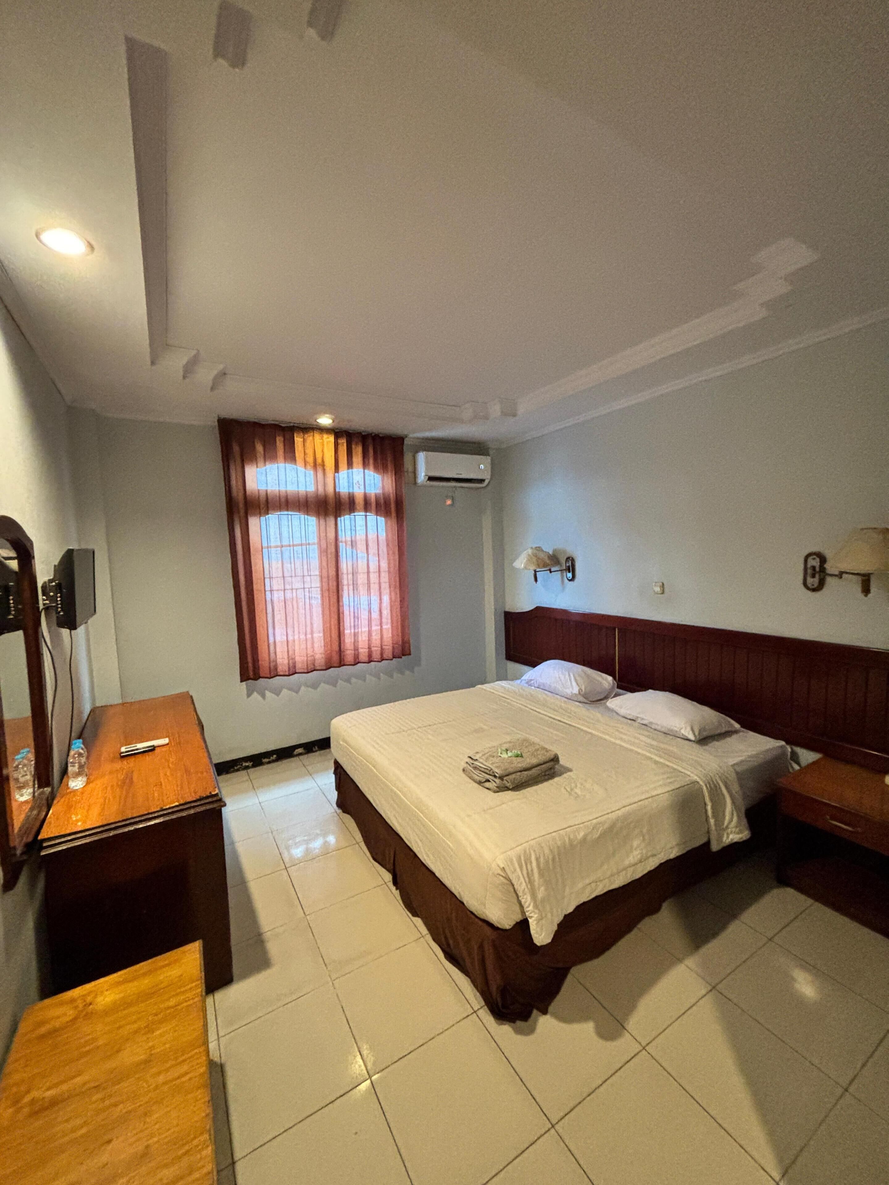 A hotel room with a bed, bedside tables, a window with curtains, an air conditioning unit, and a lamp on the wall.