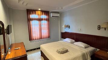 A hotel room with a bed, bedside tables, a window with curtains, an air conditioning unit, and a lamp on the wall.