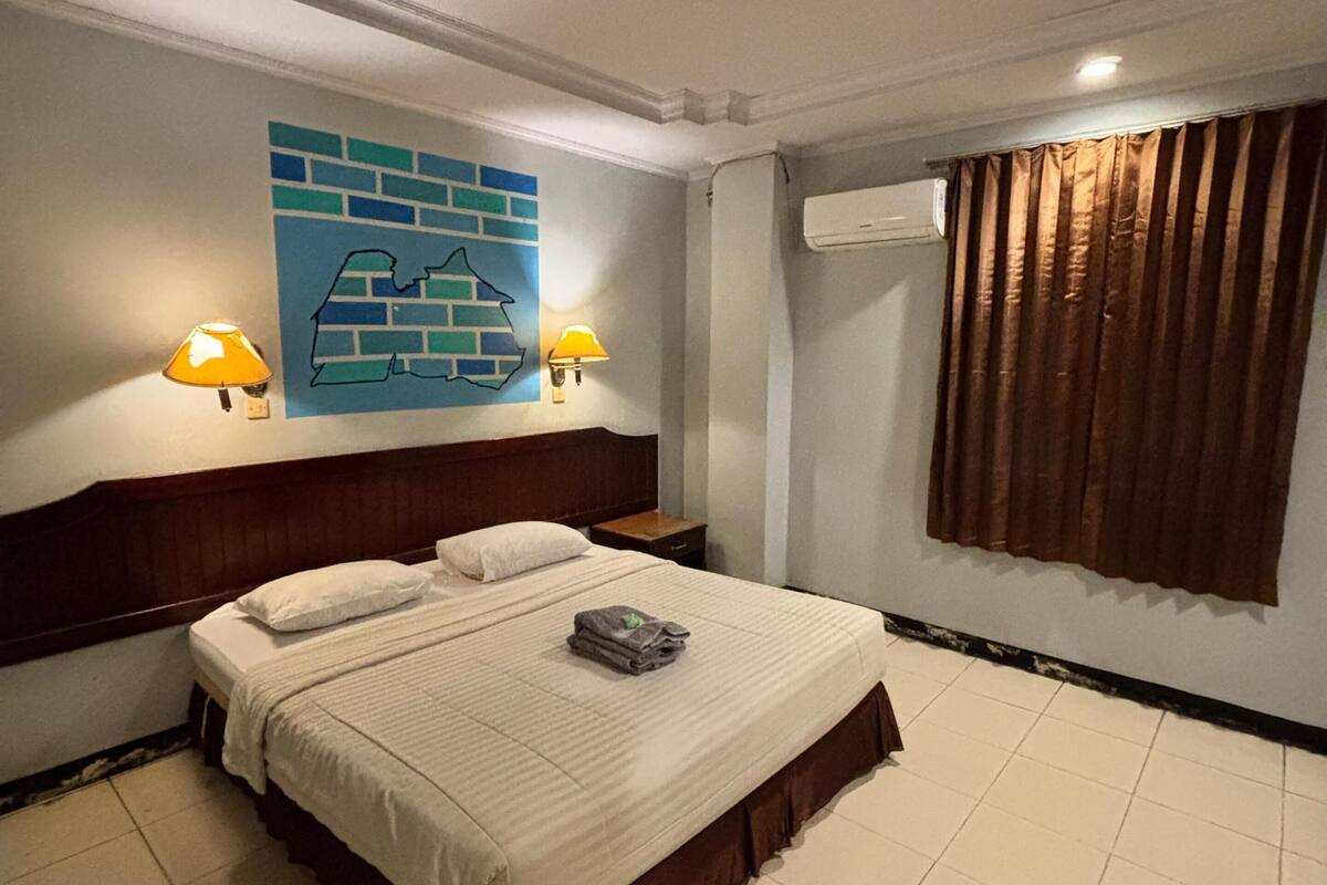 Ein Hotelzimmer mit einem Bett, zwei Wandlampen, einem Wandbild, einer Klimaanlage und einem Fenster mit Vorhängen.