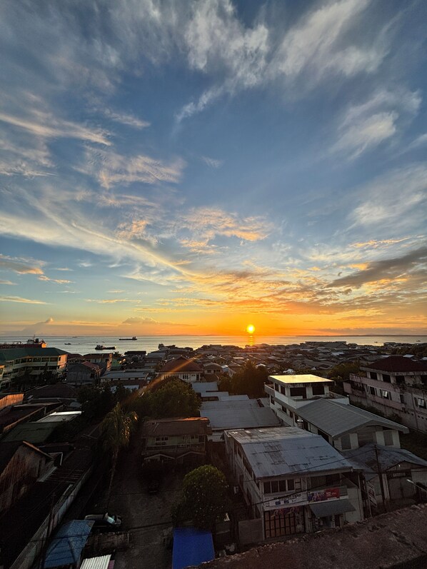 Un paisaje urbano costero al atardecer con edificios, cielo despejado y el sol cerca del horizonte.