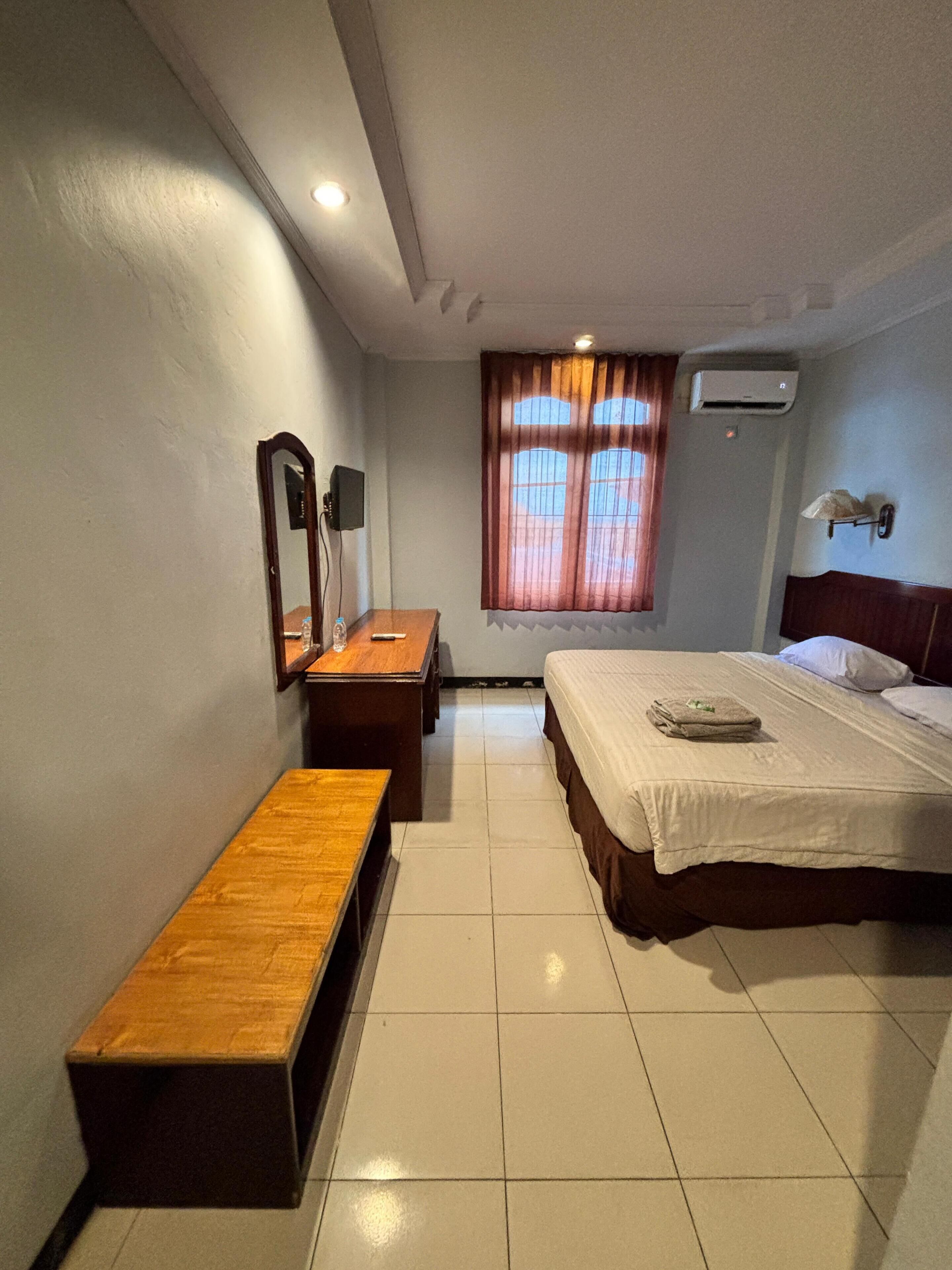 A hotel room with a bed, a desk, a bench, a mirror, an air conditioning unit, and a window with curtains.