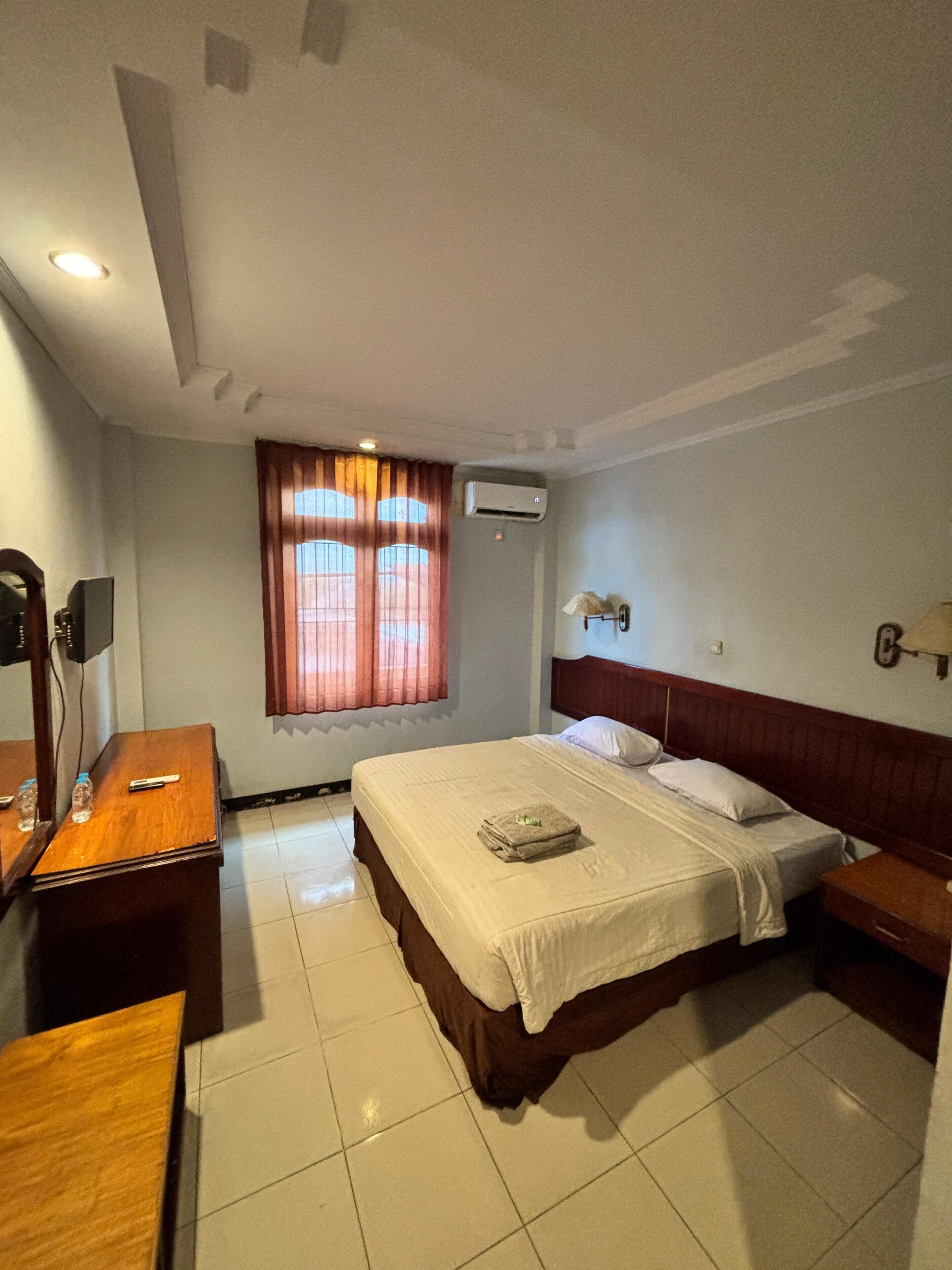 A hotel room with a bed, a desk, a chair, an air conditioning unit, and a window with curtains.