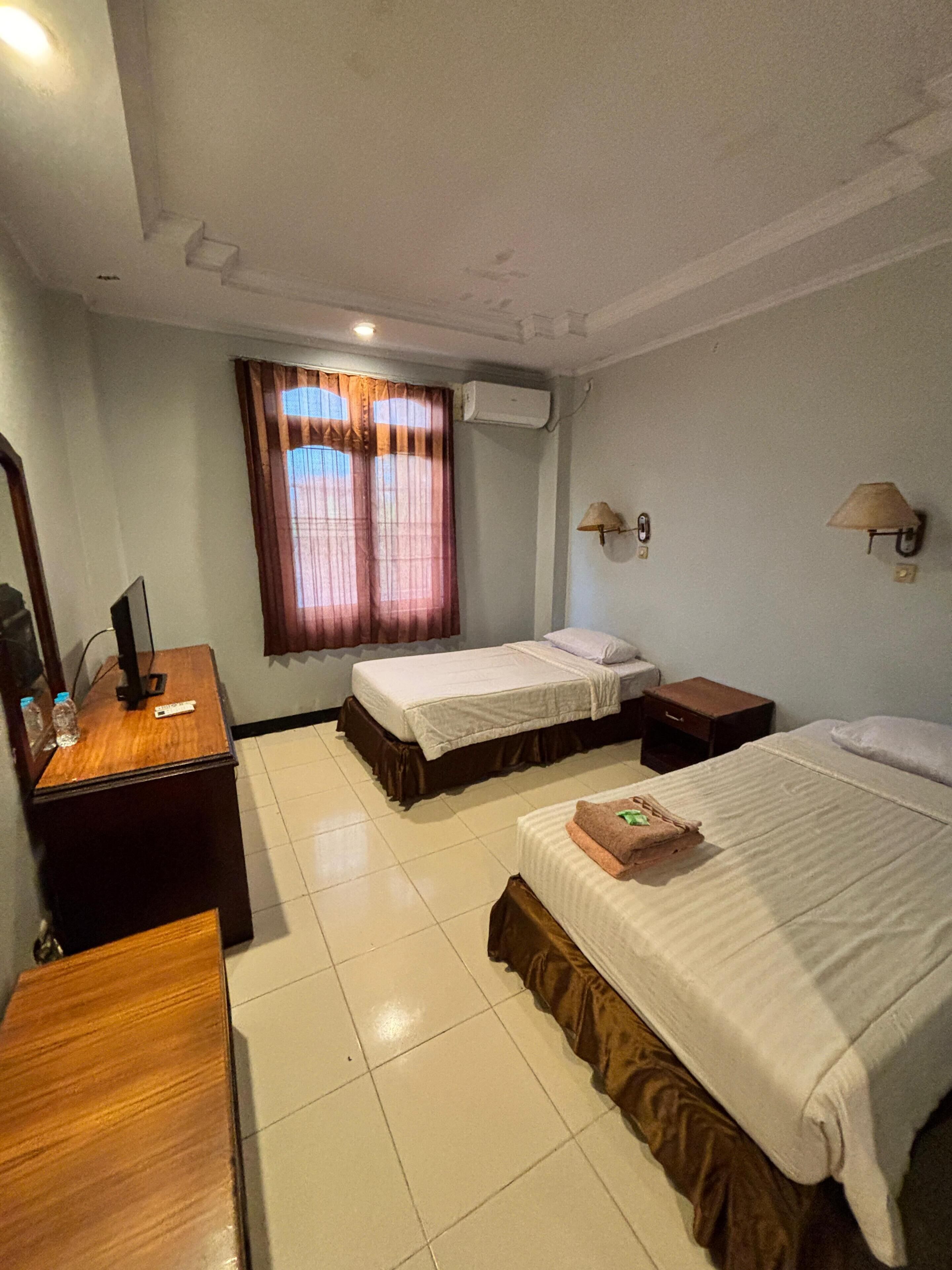 A hotel room with two beds, a desk with a computer, a mirror, a window with curtains, and an air conditioning unit.
