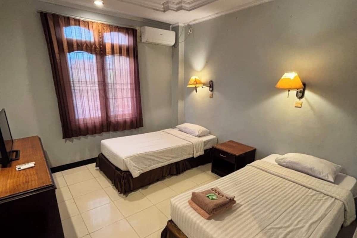 Ein Hotelzimmer mit zwei Betten, einem Schreibtisch, einem Stuhl und einem fensterbepflanzten Fenster.