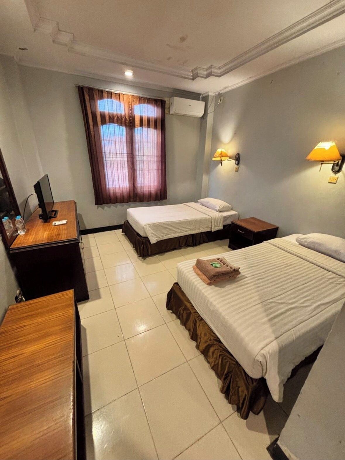A hotel room with two beds, a desk, a chair, a TV, and two wall-mounted lamps.