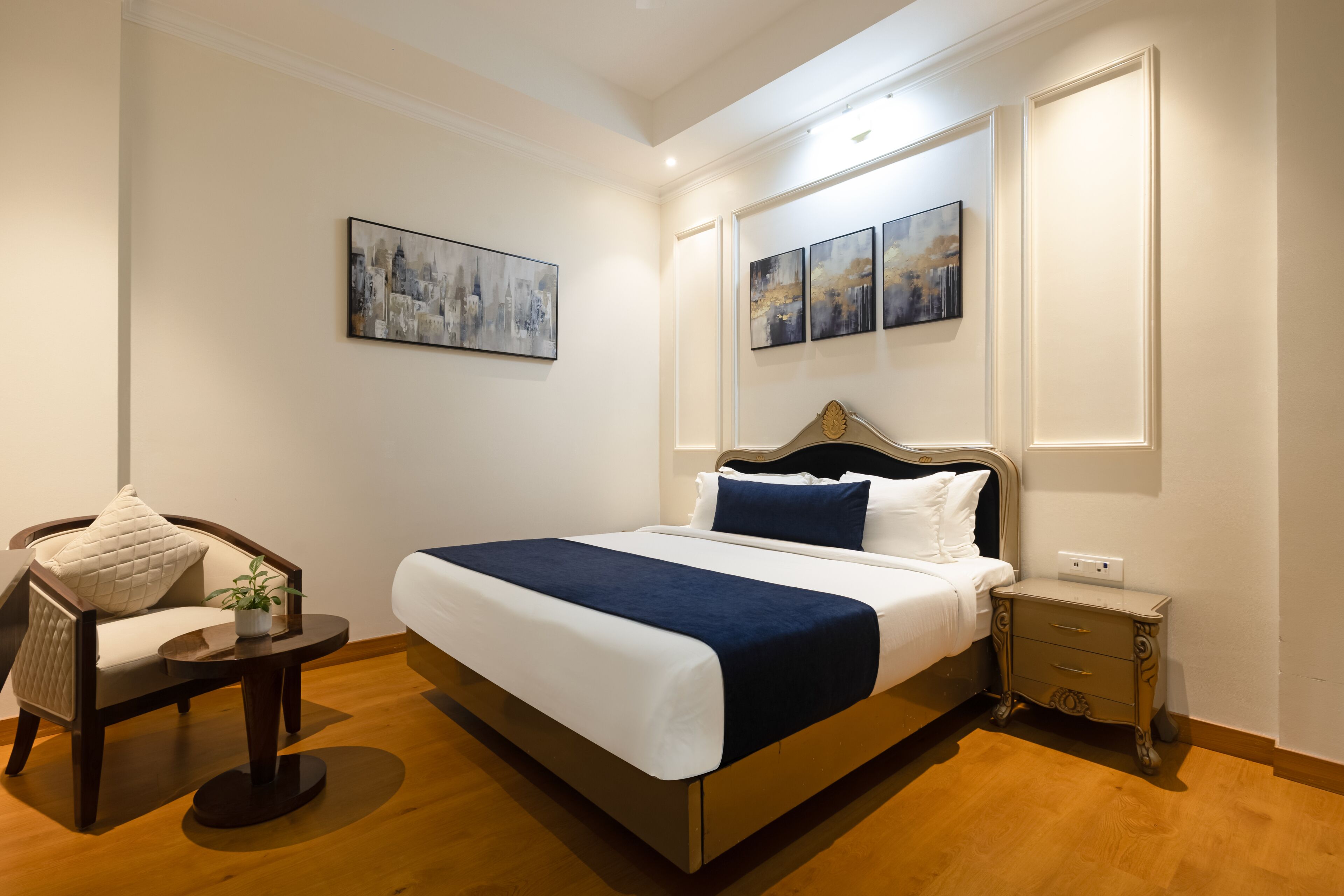 A neatly arranged bedroom with a large bed, a chair, a bedside table, and artwork on the walls.