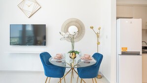 Een moderne eetruimte met een glazen tafel, blauwe stoelen, een gouden vaas met bloemen, een ronde spiegel en een flatscreen televisie aan de muur.