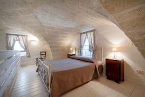 Ein Schlafzimmer mit schrägverdeckter Decke, einem Bett, einem Nachttisch, einem Sessel und einem Fenster mit Vorhängen.