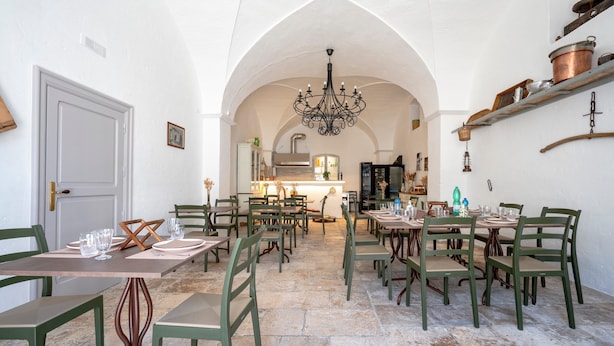 A dining area with multiple tables and chairs, a chandelier, and a kitchen in the background. - Lamapecora - Salone Apartment with Balcony, Wi-Fi and Air Conditioning (Fasano)