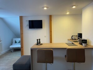 Ett modernt kök med en bänkskiva i trä, två barstolar, en kaffemaskin och en platt-TV som är monterad på väggen.