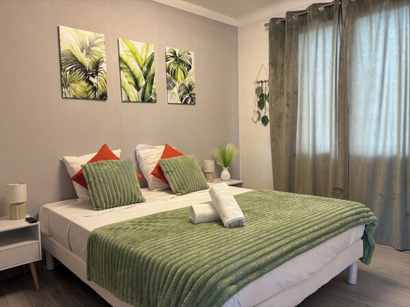 A hotel room with a bed, green and white bedding, a nightstand with a lamp, a vase with a plant, and three wall art pieces.