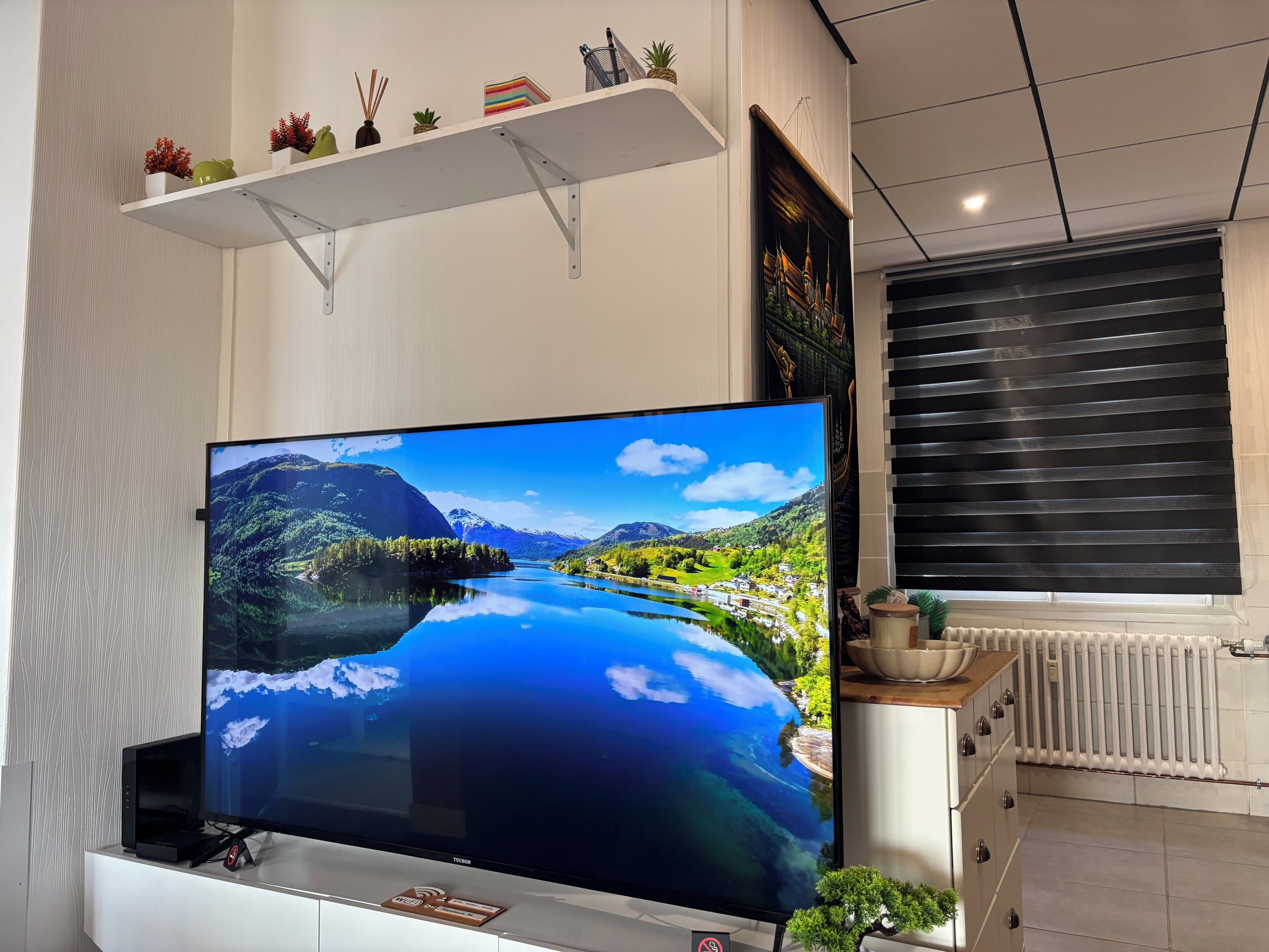 Un téléviseur à écran plat posé sur un meuble blanc, affichant une image de paysage, une étagère blanche ornée d’objets décoratifs et une fenêtre avec des stores.