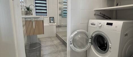 A laundry area with a washing machine, a trash bin, and a window with blinds.