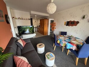 A modern living room with a sofa, a dining table set for two, and a television displaying a nature scene. - Bright 75m2 3-bedroom Apartment with a largebalcony - fully equipped (Vittel)