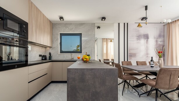Een moderne keuken met een centraal eiland, een eethoek met tafel en stoelen, en een groot raam.