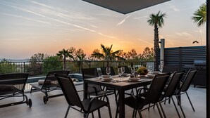 Een terras met een eettafel voor zes personen, omringd door stoelen, met uitzicht op een schilderachtig landschap met palmbomen en een zonsondergang.