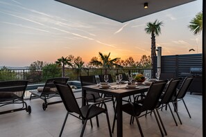 An outdoor dining area with a table set for six, surrounded by chairs, overlooking a scenic view with palm trees and a sunset. - Villa Lukante Zadarvillas (Zadar)