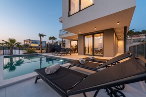 Un espace moderne aménagé au bord de la piscine, avec des chaises longues, une piscine et un bâtiment doté de grandes fenêtres et d’un balcon.