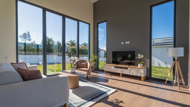 A modern living room with a sofa, TV, and large windows overlooking a green landscape. - Lotus Villas & Suites (Polygyros)