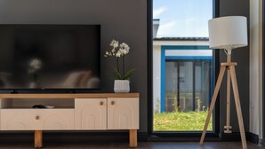A modern living room with a TV, a small white vase with flowers, a floor lamp, and a view of a blue building through a large window. - Lotus Villas & Suites (Polygyros)