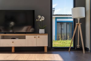 A modern living room with a TV, a small white vase with flowers, a floor lamp, and a view of a blue building through a large window.