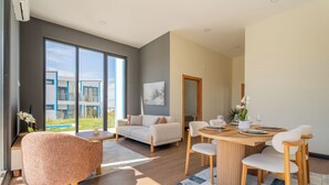 A modern living room with a sofa, a round wooden dining table with chairs, a large window with a view of a building, and a small plant on a side table. - Lotus Villas & Suites (Polygyros)