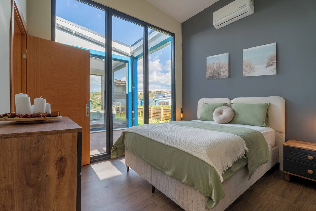 Une chambre à coucher comprenant un lit, une table de chevet, une commode en bois et une grande fenêtre donnant sur un paysage verdoyant.
