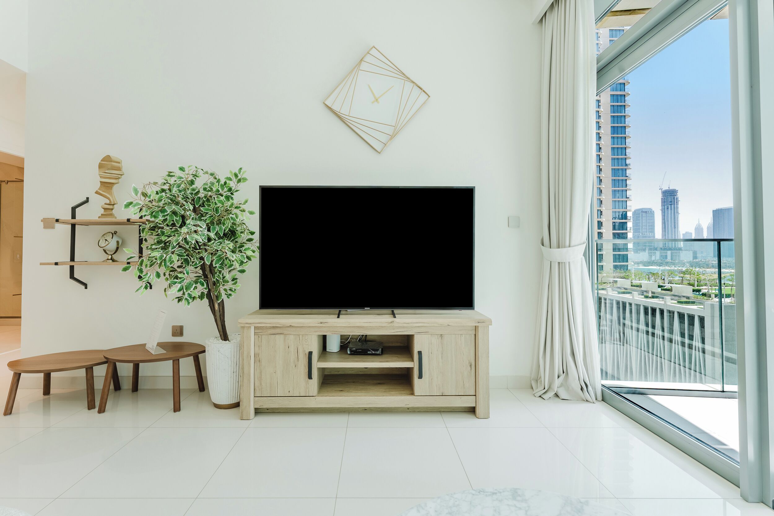 A modern living room with a flat-screen TV, wooden furniture, a potted plant, a wall clock, and a large window with a city view.