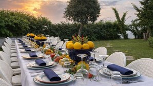 Una larga mesa de comedor dispuesta para una comida, con sillas, un centro de mesa con limones y una puesta de sol al fondo.