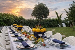 Una larga mesa de comedor dispuesta para una comida, con sillas, un centro de mesa con limones y una puesta de sol al fondo.