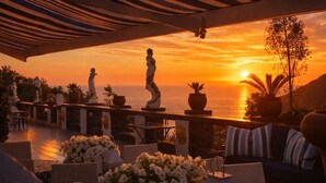 Una terraza con mesas al aire libre, vista del atardecer sobre el mar y estatuas decorativas.