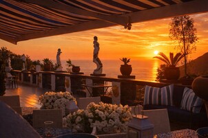 Una terraza con mesas al aire libre, vista del atardecer sobre el mar y estatuas decorativas.