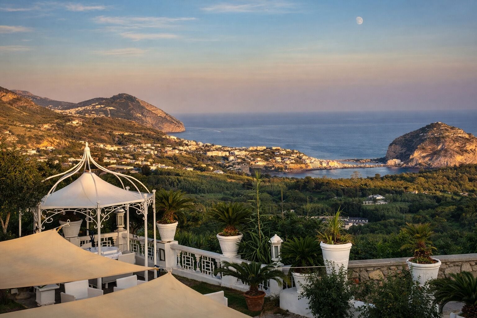 A gazebo with a white roof, surrounded by potted plants and white railings, overlooking a coastal town with mountains in the background.