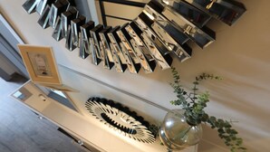 Ein modernes Interieur mit einem spiegelnden Wandkunstwerk, einer Vase mit Grünpflanzen und einem gerahmten Bild auf einer Konsole.