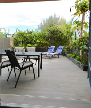 Een terras buiten met een tafel en stoelen, omgeven door weelderig groen en een uitzicht op de lucht.