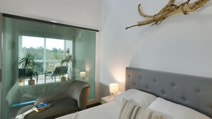 Ein modernes Schlafzimmer mit einem großen Bett, einer Glaswand, Blick ins Freie und einem Deckenventilator.