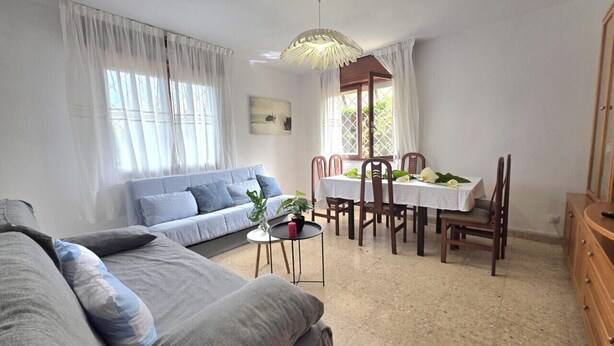 Una sala de estar con un sofá, una mesa de comedor con sillas, una pequeña mesa redonda con una planta y una lámpara colgante.
