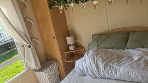 A bedroom with a bed, a nightstand, a lamp, a window with curtains, and a view of greenery outside. - 2 Bed - Sleeps 6 - Free Parking - Pets (Skegness)