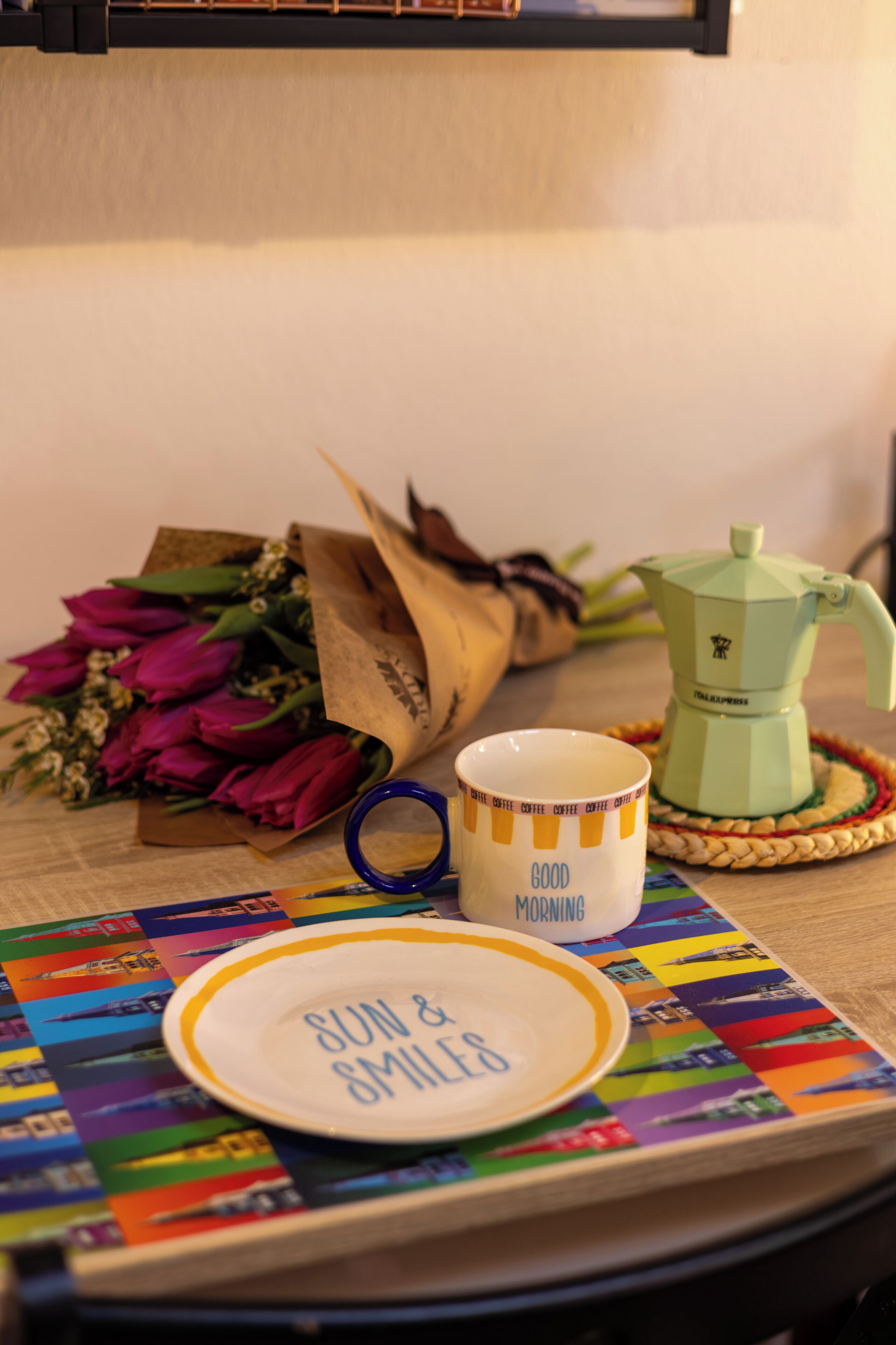A table with a coffee maker, a mug, a plate with 'SUN & SMILES' text, a colorful placemat, and a bouquet of flowers.