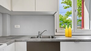 Una cocina con un lavamanos de acero inoxidable, una ventana con vistas a árboles y una puerta con picaporte rojo.