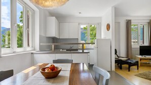 Una cocina moderna con armarios blancos, una mesa de comedor de madera con un tazón de frutas y vistas a la vegetación exterior.