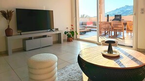 A living room with a flat-screen TV, a sofa, a dining table, and a sliding glass door leading to an outdoor patio with a barbecue. - L'Écrin d'Olmo: Charming & spacious terrace in a peaceful setting (Pietrosella)