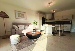 A modern living room with a sofa, a round coffee table, a floor lamp, and a kitchen area with bar stools. - L'Écrin d'Olmo: Charming & spacious terrace in a peaceful setting (Pietrosella)